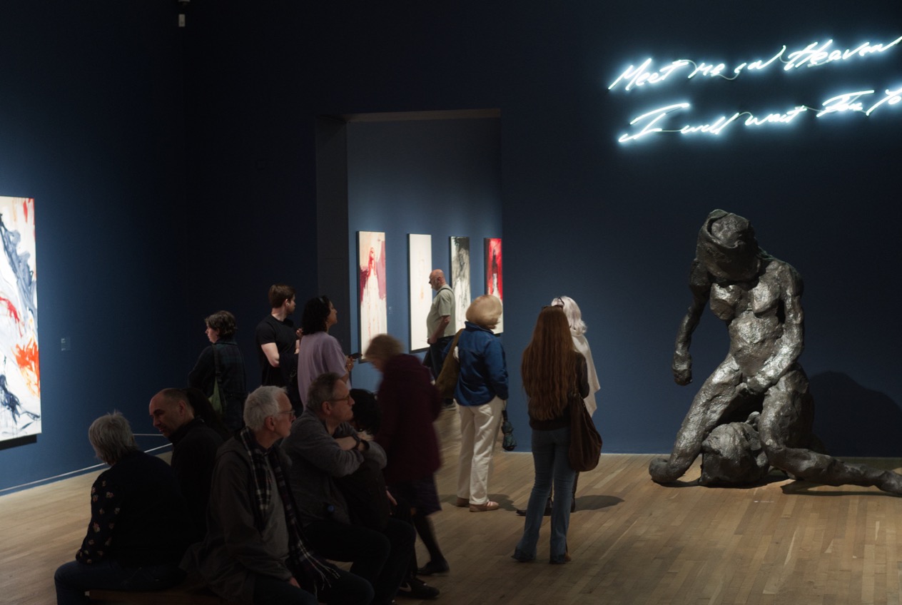 Besucher sitzen und stehen in einem dunkelblauen Saal der Tate Modern, unter einem Neonschriftzug, neben einer knienden Bronze, Mai 2026