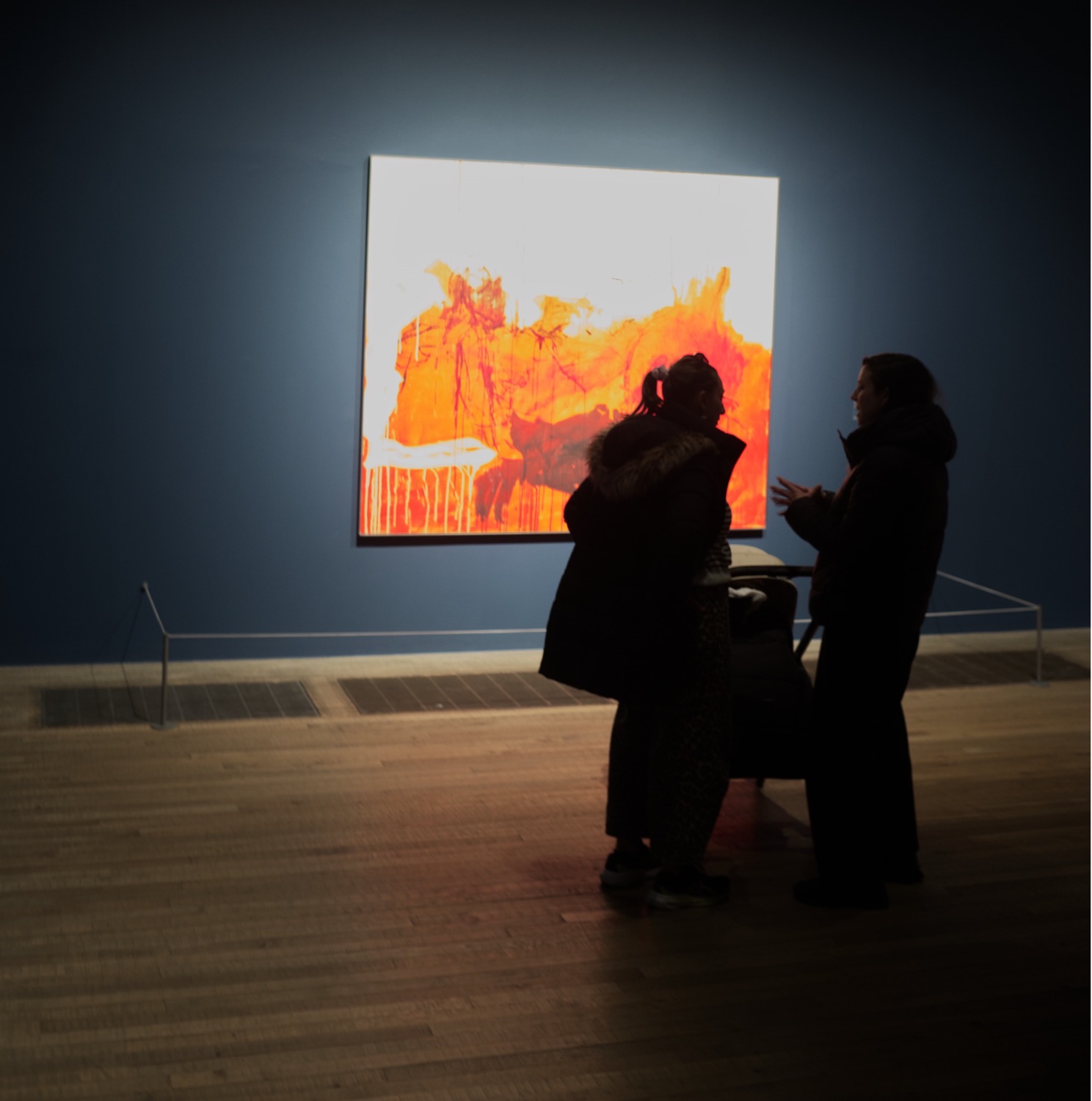 Zwei Besucher im Gespräch vor einem grossen orangen Gemälde in einem dunkelblauen Saal der Tate Modern, Mai 2026