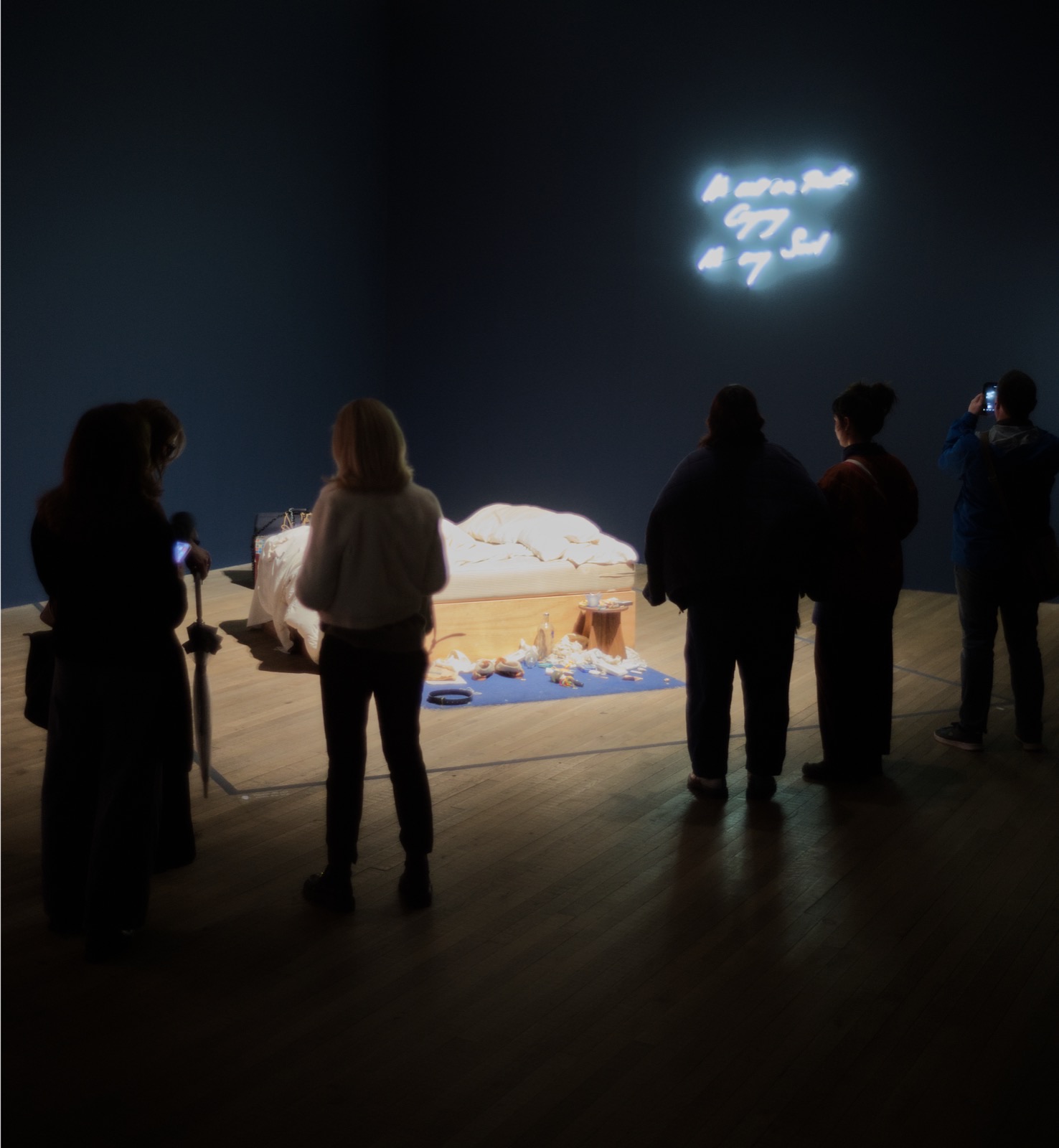 Besucher stehen vor Tracey Emins Bett in einem dunkelblauen Saal der Tate Modern, dahinter ein Neonschriftzug, Mai 2026